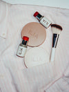 ILIA beauty products, including blush sticks, bronzer, highlighter, and a makeup brush, placed on a pink striped shirt with some items in a pocket.