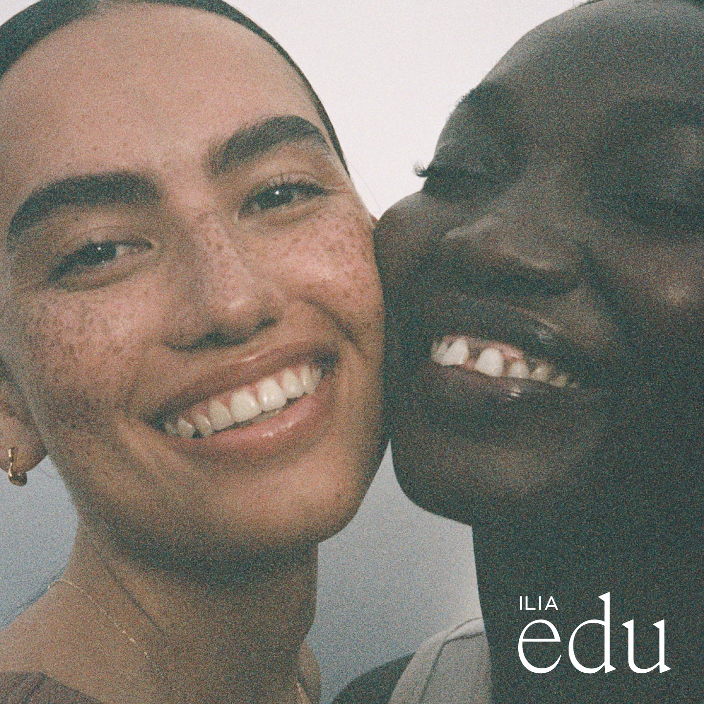 Two smiling models with radiant skin embracing closely, promoting natural beauty and edu program by ILIA.
